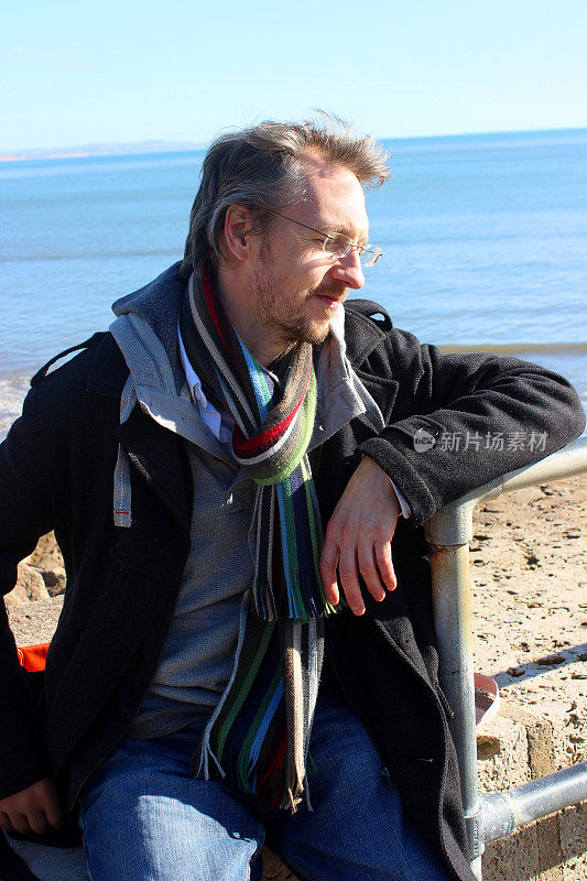一个男人穿着冬天的大衣/围巾坐在沙滩上